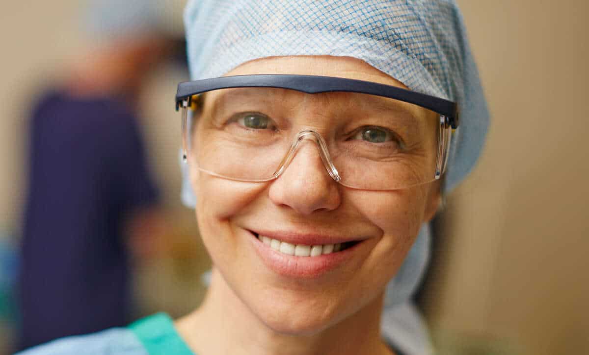 Surgical nurse with protective eyewear in operating room, smiling, wearing scrubs and surgical cap, healthcare professional, medical team, patient care, hospital surgery, clinical environment, surgical staff, medical profession.